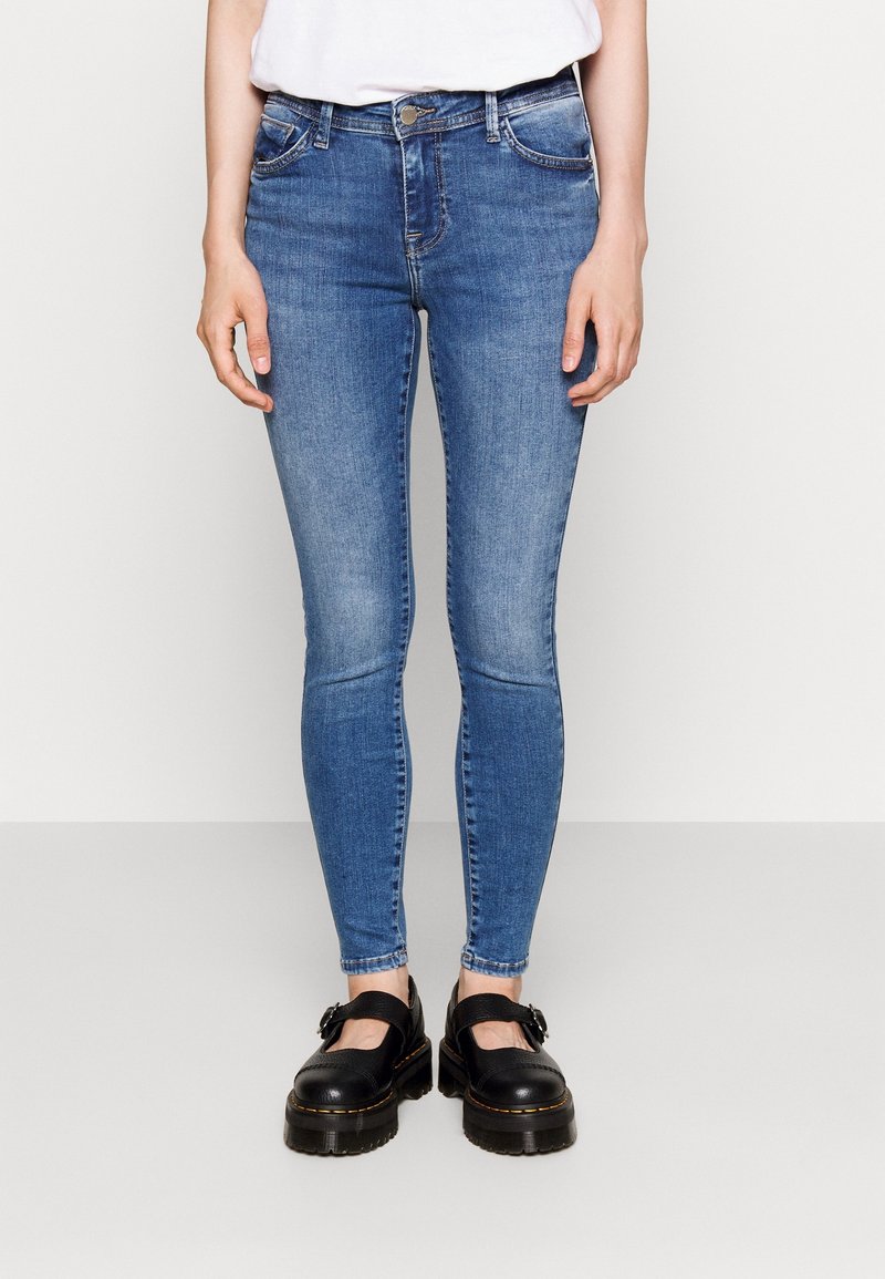 Skinny jeans in medium blue denim with a faded design. Features a five-pocket style, belt loops, and a button closure. Worn with black shoes.
