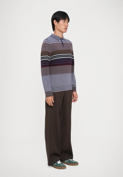 Langermet polo skjorte med horisontale striper i brune, lilla og blå nyanser; kombinert med vide brune bukser og grønne sneakers.