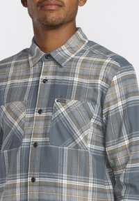 Chemise en flanelle à carreaux grise, beige et blanche. Comprend deux poches poitrine et une fermeture à boutons. Texture douce avec un design de col classique.