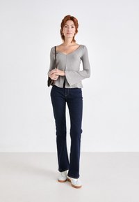 Maglione grigio a coste con maniche lunghe, collo a V e polsini svasati, abbinato a jeans svasati blu scuro e sneaker bianche con suole marroni.