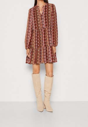 Kvinde iført en langærmet, mønstret kjole over knæene i varme toner, kombineret med beige ruskindsstøvler til knæet mod en plain lys baggrund.