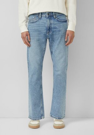 Personne portant un jean droit bleu clair, des baskets blanches et un haut à manches longues crème, debout devant un fond uni.