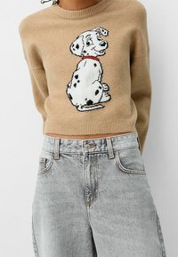 Beiger Pullover mit einem Dalmatiner-Grafikdesign, rundem Ausschnitt und geripptem Saum, kombiniert mit hoch taillierten hellblauen Jeans.