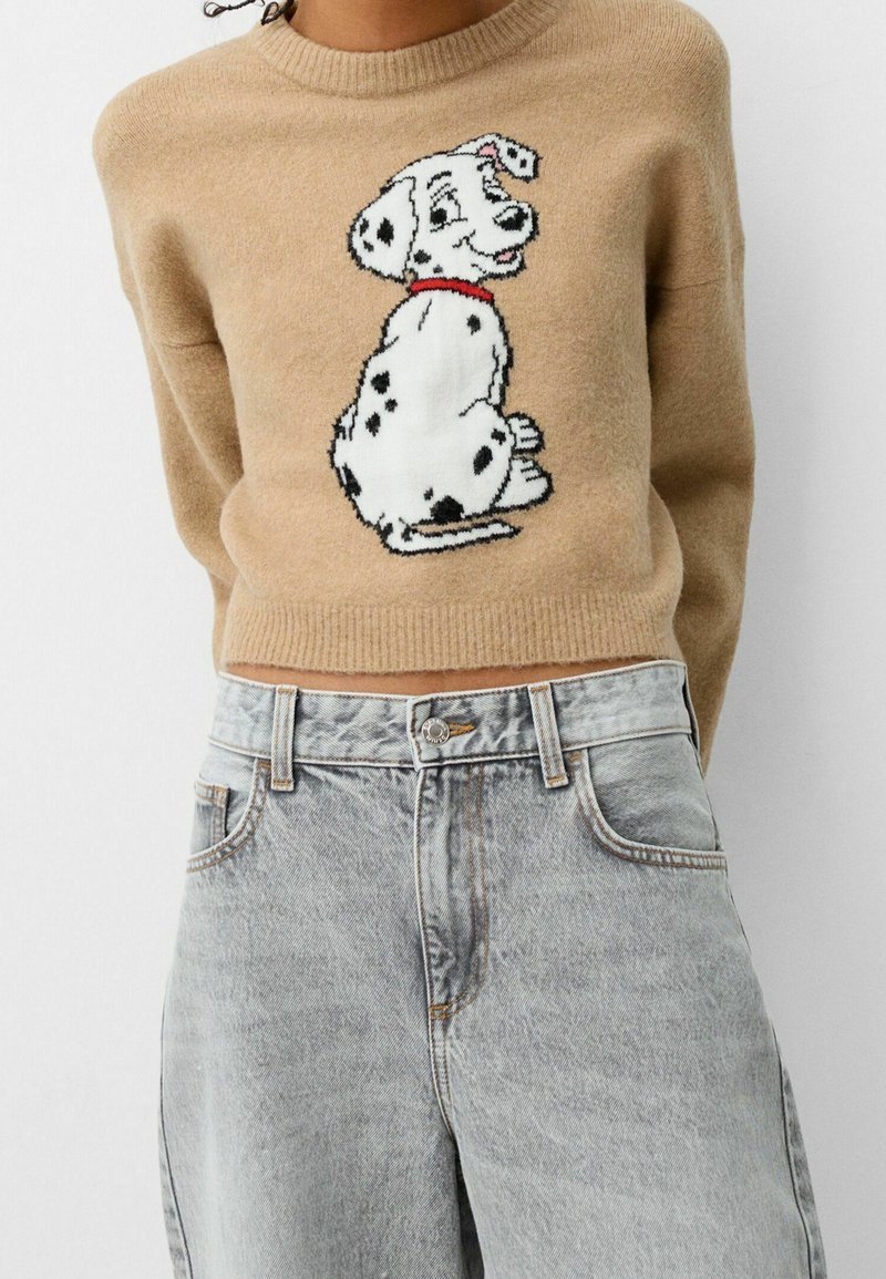 Beiger Pullover mit einem Dalmatiner-Grafikdesign, rundem Ausschnitt und geripptem Saum, kombiniert mit hoch taillierten hellblauen Jeans.