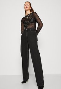 Γυναίκα φορώντας μαύρη διαφανή μπλούζα με μακριά μανίκια και κεντήματα, μαύρα ψηλόμεσα φαρδιά τζιν, μαύρες μυτερές γόβες και στρωτές αλυσίδες, στέκεται με αυτοπεποίθηση.