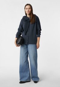 Granatowa koszula na guziki z teksturowanym wełnianym szalikiem, połączona z szerokimi, jasnoniebieskimi dżinsami i małą czarną torebką.