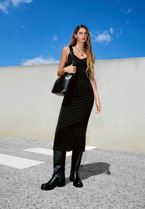 Große Frau mit langen Haaren, die ein schwarzes ärmelloses Kleid und schwarze knielange Stiefel trägt. Sie hält eine schwarze Umhängetasche und steht im Freien.