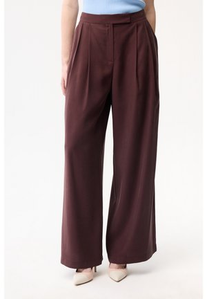 Donna che indossa pantaloni marroni a vita alta e gamba larga abbinati a una canotta azzurro chiaro e tacchi beige, in piedi davanti a uno sfondo uniforme.