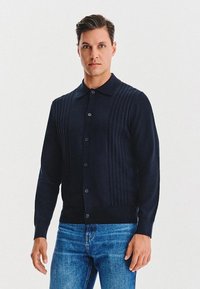 Mężczyzna z krótkimi ciemnymi włosami, noszący zapinaną granatową kardigan z pionowym prążkowaniem i niebieskie dżinsy, stoi na tle gładkiej białej ściany.