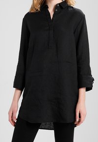 Zwart linnen tuniek met een kraag, een kwart-knopsluiting en lange mouwen. Heeft een ontspannen pasvorm en een lichtgebogen zoom.