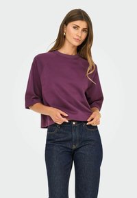 Femme aux longs cheveux portant un sweatshirt court à manches courtes violet et un jean bleu foncé, accessoirisée avec des boucles d'oreilles en or, un bracelet et une bague.