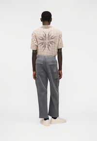 Καφέ καρό κοντομάνικο πουκάμισο με μεγάλο μοτίβο ηλιακής ακτίνας στην πλάτη, σε συνδυασμό με γκρι παντελόνι cargo και μπεζ αθλητικά παπούτσια.