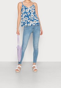 Haut sans manches floral bleu avec des fleurs blanches, jean skinny en denim clair, sandales blanches et sac fourre-tout en maille lavande. Tissu doux, coupe décontractée.