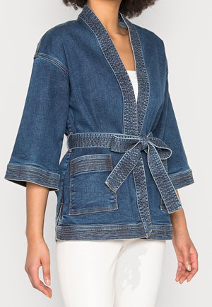 Personne portant une veste kimono en denim bleu avec des manches larges, des poches avant et une ceinture en tissu nouée, sur un pantalon blanc.
