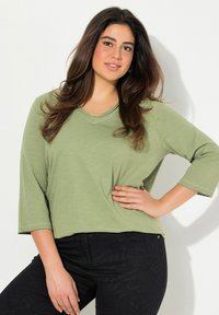 Světlý zelený top s dlouhým rukávem, zaobleným výstřihem ve tvaru V a prošíváním, doplněný černými vzorovanými džíny. Modelka se pózuje s rukou na bocích.