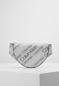 Calvin Klein-väska i vitt syntetmaterial, med en rundad klaffdesign. Svart logotext tryckt över ytan, med kontrasterande sömmar.