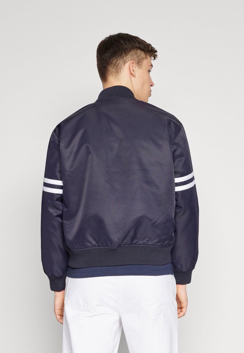 Un jeune homme de dos portant une veste bomber navy avec des rayures blanches sur les manches et un pantalon blanc, devant un fond uni.
