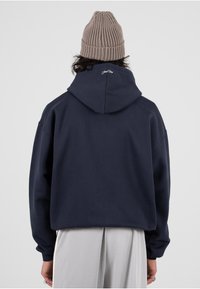 Sudadera oversized azul marino con capucha con cordón y puños acanalados. Combinada con pantalones gris claro. Cuenta con un sutil logotipo en la capucha.