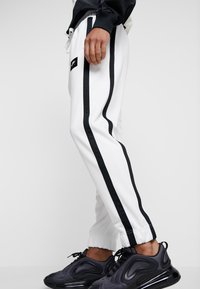 Vita sportbyxor med svarta sidosträck, tillverkade av en slät tyg, med elastisk midja och fotledsmuddar. Framträdande svart logotypmärke.