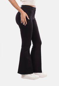 Pantaloni neri a zampa d'elefante in un tessuto morbido e elasticizzato. Presentano una chiusura con bottone, un design aderente fino al ginocchio e poi si allargano. Indossati con sneakers bianche.