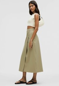 En ljusbeige kort topp i kombination med en lång, flödig, khaki A-linje kjol. Kjolen har veck och en slät textur. Svarta platta sandaler används.