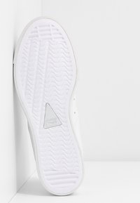 Witte sneakerzool met een gestructureerd, golvend oppervlak. Bevat een driehoekig grijs logoaccent in het midden. Plat van vorm met een minimalistisch ontwerp.