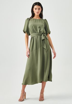 Olive green, knee-length dress made of textured fabric, featuring puff sleeves, a tied waist, and an asymmetrical hem. Heeled sandals.