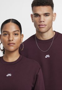 Sweatshirts bordô com gola redonda e logótipo branco da Nike SB. Um modelo usa brincos de argola, o outro tem um colar de corrente prateada.