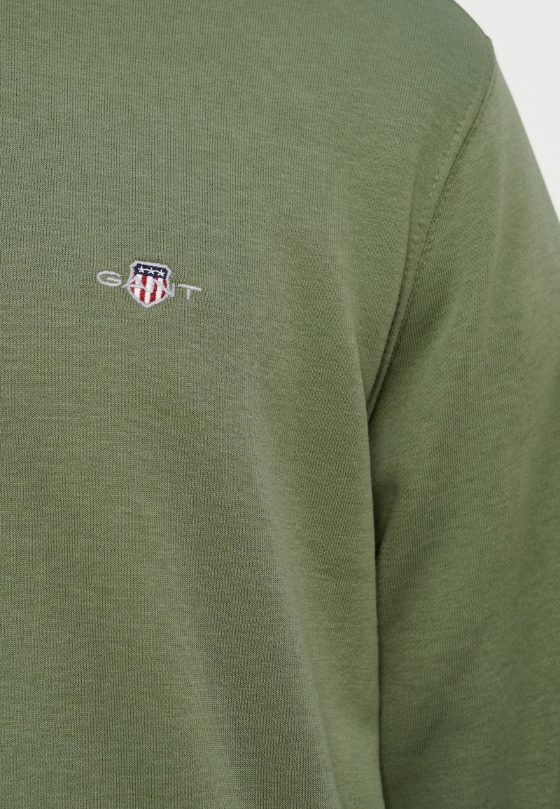 Camiseta de manga larga color verde oliva, hecha de algodón, con un pequeño logo bordado de GANT en el pecho. Textura suave y ajuste relajado.