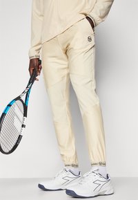 Kremfargede treningbukser med smale ben og elastiske mansjetter, parret med hvite sneakers, holder en tennisskay.