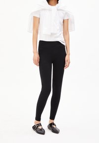 Svarta leggings med en slät, åtsittande design och hög midja, kombinerade med en vit topp som accentueras av en drapera sjal. Svarta platta skor fulländar looken.