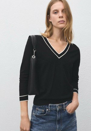 Pull noir à manches longues avec un col en V et des poignets blancs, associé à un short en denim. Mannequin portant un sac à main noir sur son épaule.