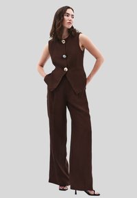 Hnědá upravená vesta s vysokým límcem, zdobená dekorativními knoflíky, v kombinaci s šírokými kalhotami a černými sandály na podpatku.