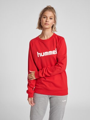 Ung kvinde stående iført en rød sweatshirt med "hummel" logo og lysegrå joggingbukser mod en ensartet lys baggrund.