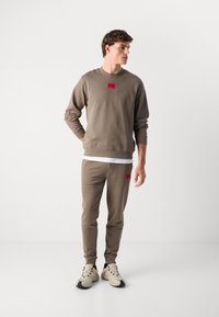 Felpa marrone con pantaloni coordinati, entrambi con un patch logo rosso. Il materiale è morbido con una finitura strutturata, con polsini e vita a costine.