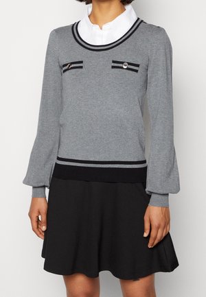 Haut tricot gris avec un col rond et des rayures noires, comportant des poches sur le devant et une jupe noire, créant un look superposé. Texture douce.