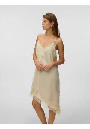 Femeie purtând o rochie tip camisă din satin bej cu tiv din dantelă, bretele subțiri și tiv asimetric, stând în picioare pe un fundal simplu, deschis.