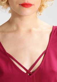 Haut bordeaux avec un décolleté en V présentant des bretelles croisées au niveau du cou. Texture lisse et tissu léger. Vue en gros plan.