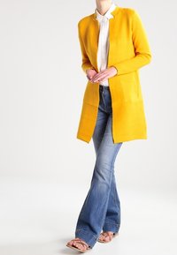 Dlouhý žlutý kardigan z texturované pleteniny, s otevřeným předním dílem, kombinovaný s rozšířenými modrými džínami a sandály.