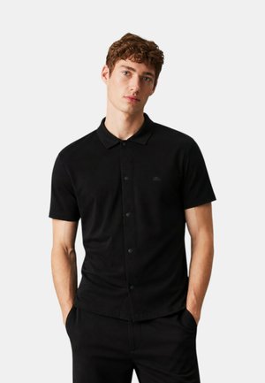 Giovane uomo con capelli ricci che indossa una camicia nera a maniche corte con bottoni e pantaloni neri, mani nelle tasche, sfondo neutro.