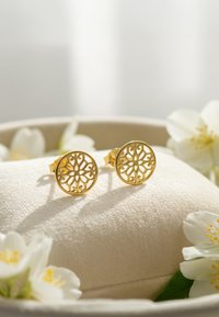 Guld filigran studs med blomster mønstre, cirkulær form og struktureret overflade. Præsenteret på en blød beige pude omgivet af hvide blomster.