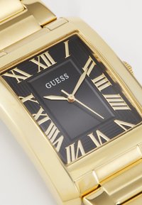 Rektangulær guldur med sort urflade, romerske tal som markører og guld-urhænder. Har en struktureret bezel og et guldmetalarmbånd.