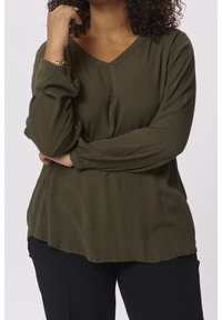 Blouse vert olive à manches longues avec un col en V, en tissu texturé et à coupe décontractée ; présente des poignets à boutons et un ourlet incurvé. Assortie à un pantalon noir.
