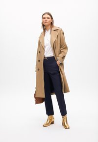 Trench coat beige con grandi bottoni, camicia bianca con un motivo sottile, pantaloni blu navy, stivaletti con tacco dorati e borsa marrone.