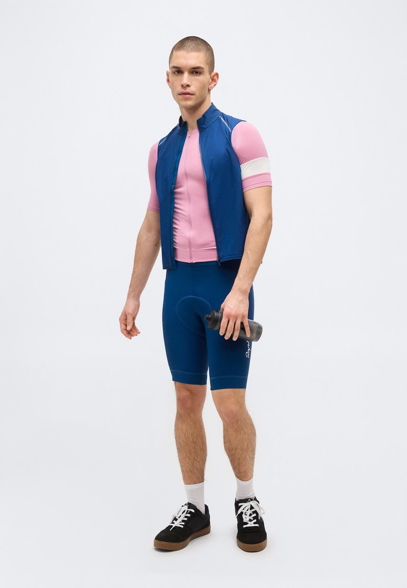 Young man in blue cycling shorts, pink short-sleeve jersey, and blue vest holds a water bottle, standing against a plain background.