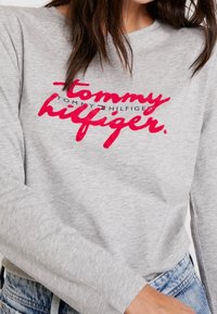 Szary t-shirt z długim rękawem z wyraźnym czerwoną, kursywną logo. Miękka tkanina, luźny krój, okrągły dekolt i ściągacze przy mankietach.