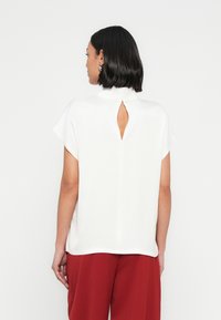 Valkoinen, lyhythihainen paita, jossa korkea kaulus ja selän avainreikäyksityiskohta. Sileä pinta, rento istuvuus, yhdistettynä punaisiin housuihin.