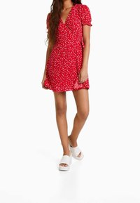 Vestido rojo con estampado de flores blancas, escote en V, mangas abullonadas y un dobladillo fluido. Combinado con sandalias de plataforma blancas.