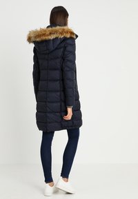 Mörkblå puffjacka med fuskpälsfoder vid huvan, med ett vadderat mönster, långa ärmar och knälång längd.