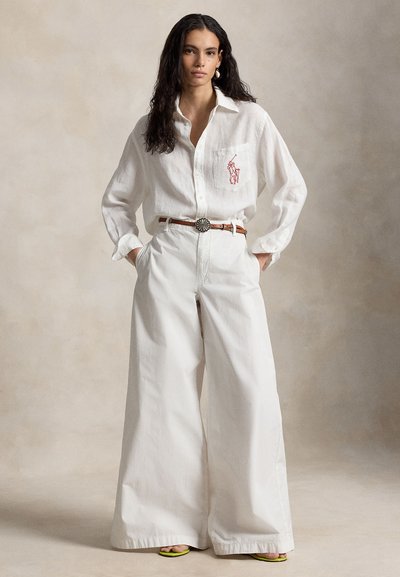 Vrouw met lang donker haar draagt een los wit overhemd met rood polo-logo, wijd uitlopende witte broek, bruine riem en limegroene sandalen.